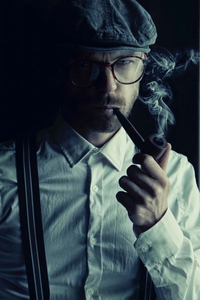 Hombre con boina y gafas de vista fumando con una pipa , vestido con camisa blanca y tirantes