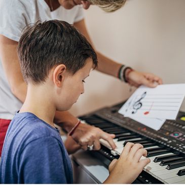piano teacher pointing to keyboard and holding colored music note flashcard, while boy plays piano