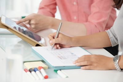 Faceless shot of contemporary colleagues using tablet while writing on clipboard sitting at table