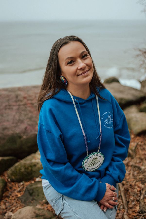 Woman in a blue hoodie sitting outdoors by the water and rocks, smiling gently.