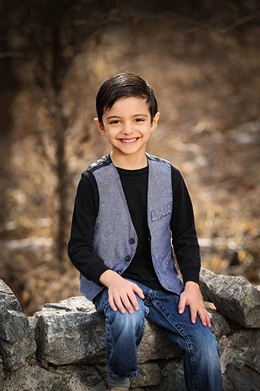 A young boy with a cheerful smile sitting on a stone wall outdoors.
