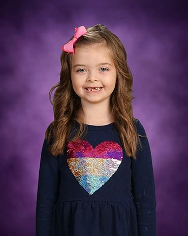 Young girl with a pink bow and sequin heart shirt smiles against a purple background.
