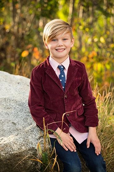 Smiling young boy in a burgundy blazer sitting outdoors on a rock.