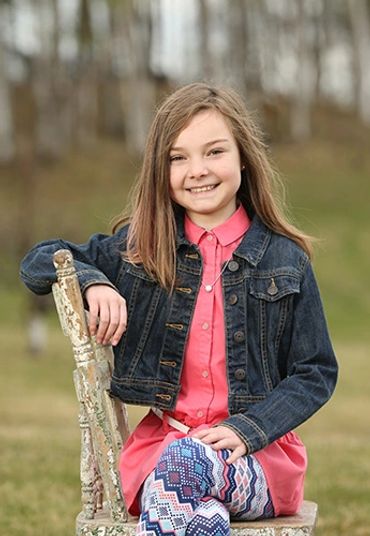 A smiling girl in a denim jacket sitting on a weathered chair outdoors.