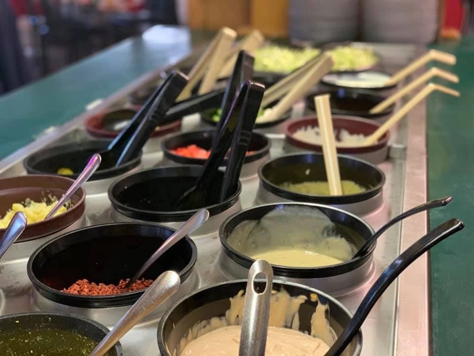 A variety of sauces and toppings with serving spoons on a buffet line.