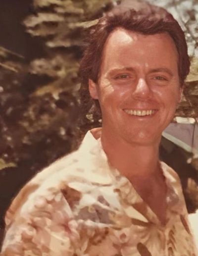 A smiling man in a patterned shirt outdoors with trees in the background.