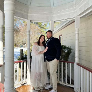 Couple posing on a porch with greenery and snow in the background.