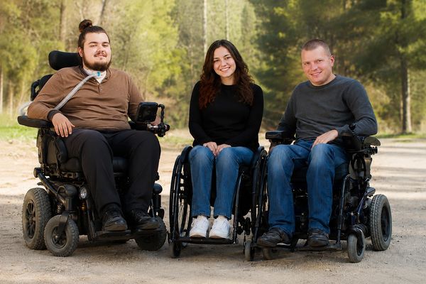 Three smiling people in wheelchairs outdoors on a sunny day.