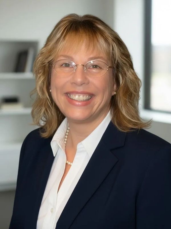 Professional woman smiling in business attire with glasses and pearls.