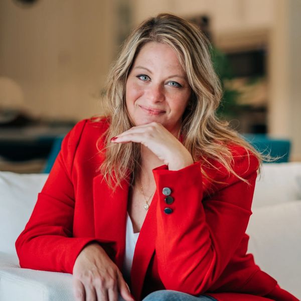 Woman in a red blazer smiling while seated on a white couch.