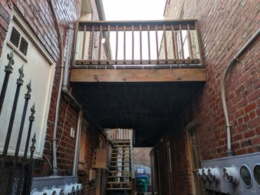 Alleyway with brick walls, utility meters, and a wooden balcony above.