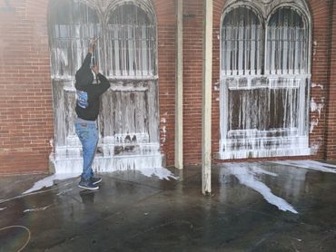 Person power washing a brick wall with wooden doors covered in white foam.