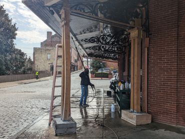 Man power washing a covered sidewalk in a historic urban area on a sunny day.