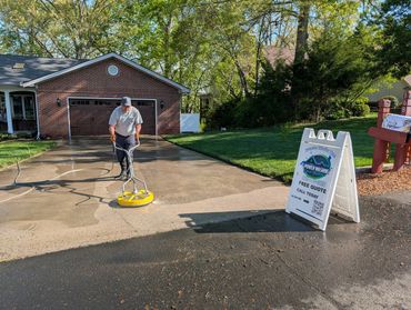 Man power washing a driveway outside a brick house.