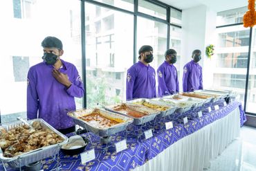 Caterers in purple uniforms stand behind a buffet table with various dishes.