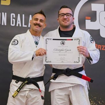 Two men in Brazilian Jiu-Jitsu uniforms at a graduation gala holding a black belt certificate.