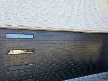 Modern black garage door with four horizontal windows on a white brick house.