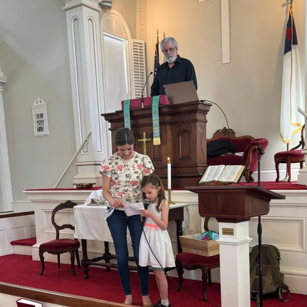 Young girl and woman reading in church with man at pulpit.