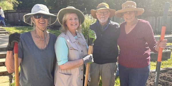 Four smiling adults in hats holding gardening tools outdoors on a sunny day.