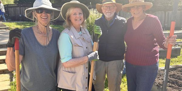 Four smiling adults in hats holding gardening tools outdoors on a sunny day.