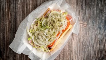 Sub sandwich loaded with onions, lettuce, tomato, and seasoned with herbs on a wooden table.