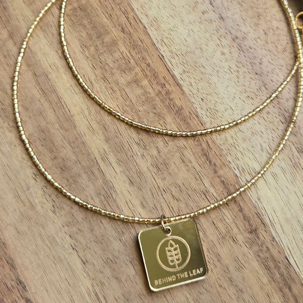 Gold beaded necklace with a square pendant engraved 'Behind The Leaf'.