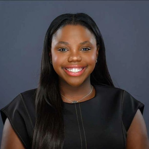 A smiling Black woman with long hair wearing a black shirt in front of a gray/blue background.