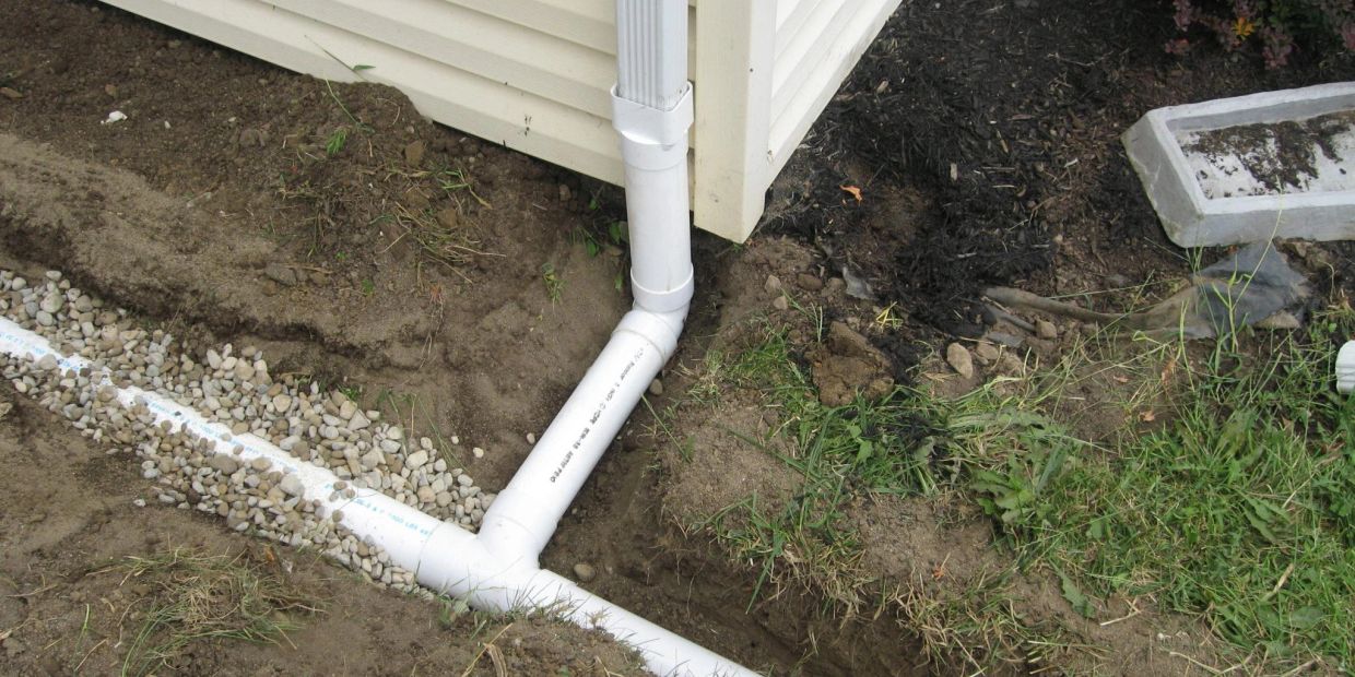 White PVC pipe drainage system installed at a house corner with gravel and soil.