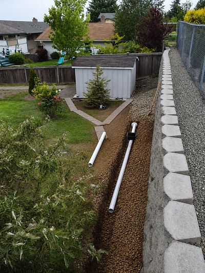 Backyard drainage system installation with white pipes and gravel next to a retaining wall.
