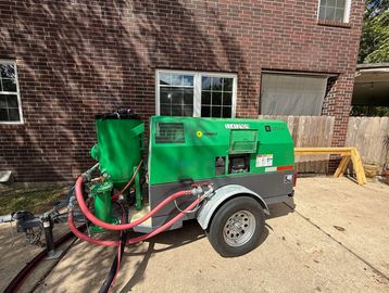 Green industrial equipment trailer with hoses, parked outside a brick building.