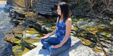 Woman meditating by the rocky water edge.