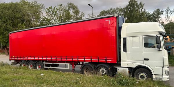 White truck with a large red trailer parked near greenery under a cloudy sky.