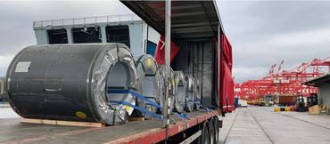 Large steel coils secured on a truck at an industrial port with red cranes in the background.