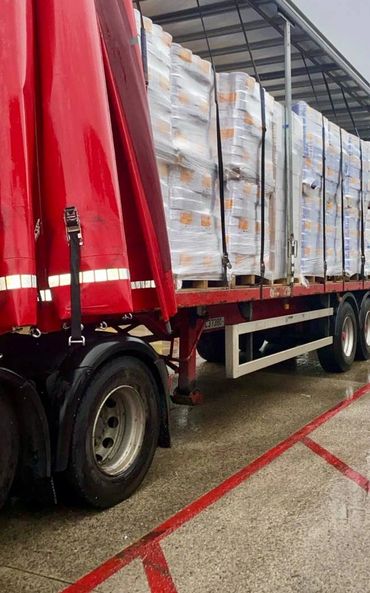 Open trailer truck loaded with wrapped pallets secured by black straps.