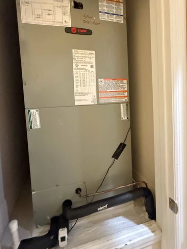 A Trane HVAC unit installed in a small closet space with visible pipes and wiring.
