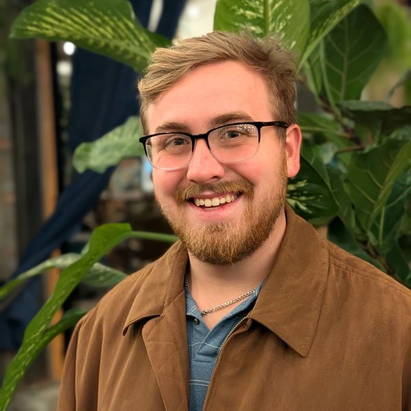 Smiling man with glasses and beard in a brown jacket, standing indoors with plants.