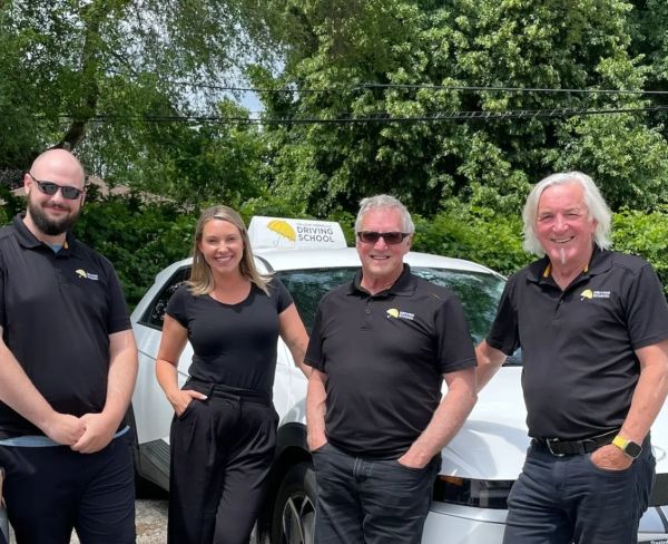Four driving school instructors standing in front of a white car with a driving school sign.