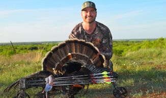 Hunter posing with a wild turkey and a bow in a grassy field.