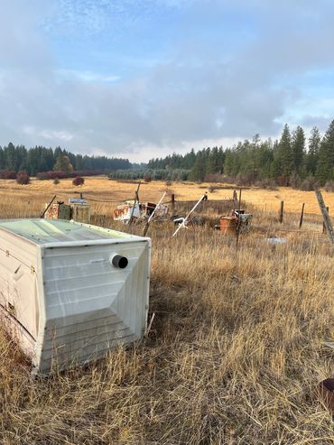 Rusty old appliances and a toppled container in a dry field with trees in the background.