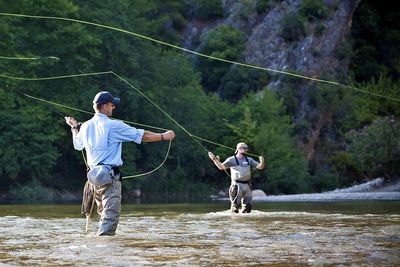 Two men fly fishing in a river surrounded by forest and cliffs.