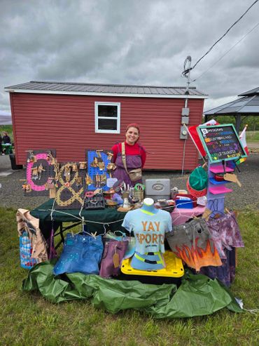 Vendor at an outdoor market with handmade crafts and clothing on display.