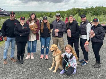Group photo of people outdoors with a dog, holding bottles and flowers under cloudy sky.