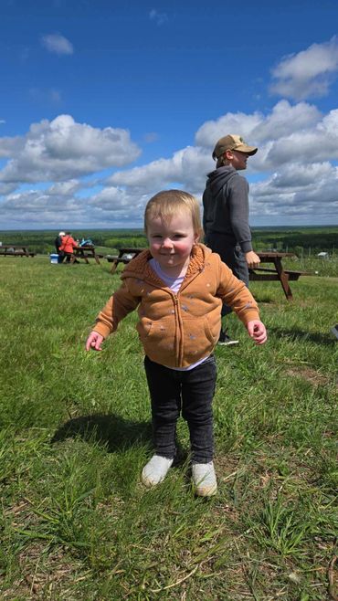 A toddler in an orange jacket stands on grass under a blue sky with scattered clouds.