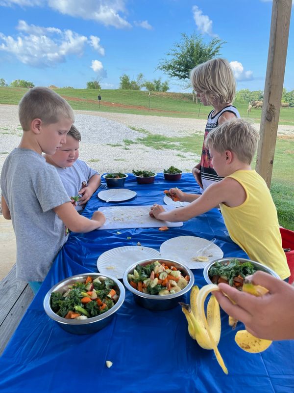Children preparing vegetables outdoors on a blue tablecloth under a clear sky.