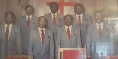 Seven men in matching suits standing in front of a cross.