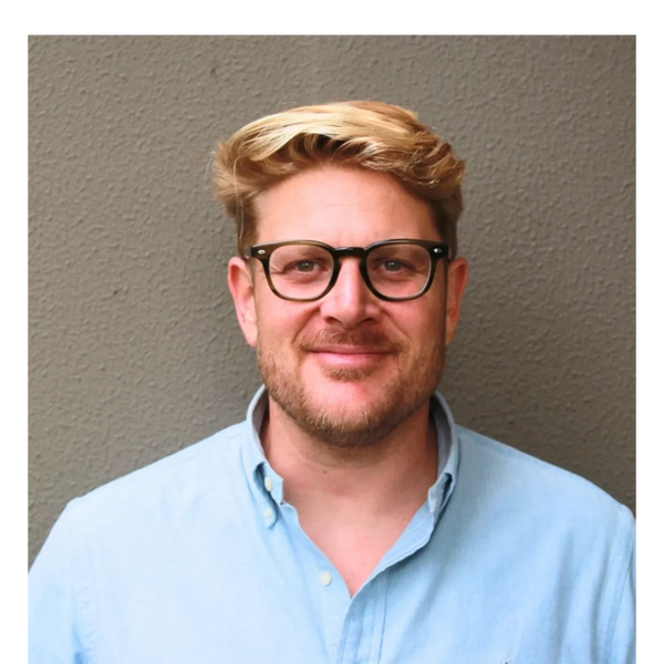 Smiling man with glasses and blond hair wearing a light blue shirt.