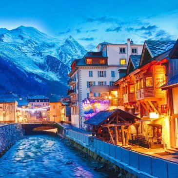 Cidade charmosa com casas iluminadas e montanhas nevadas ao fundo.