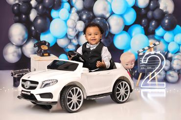 A toddler in a toy car at a blue and black balloon-themed birthday party.