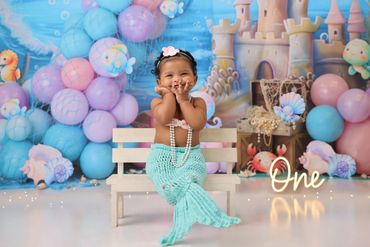 Adorable baby dressed as a mermaid sitting on a bench with ocean-themed decorations.