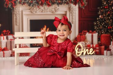 Smiling baby girl in a red dress celebrating her first birthday with festive decorations.